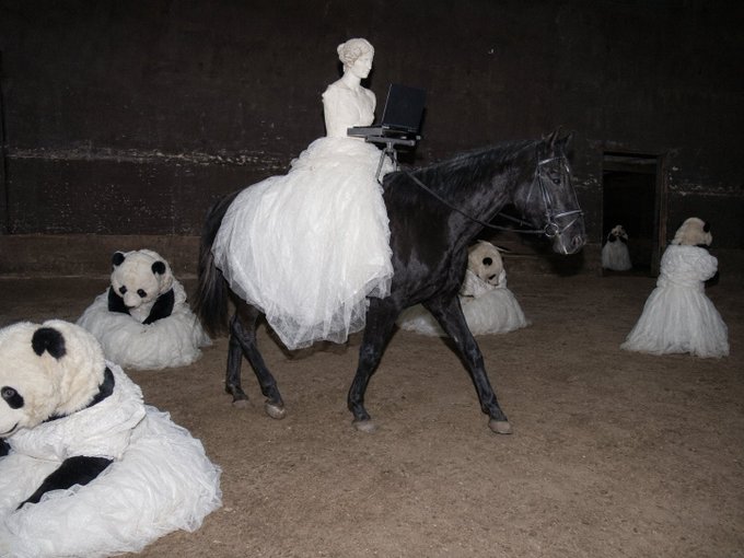 A surreal art photograph featuring a woman in a white ball gown riding a black horse while using a laptop, surrounded by figures in white dresses with panda and teddy bear heads in a dimly lit barn-like setting.