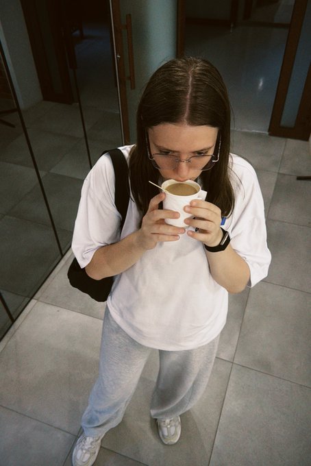 A young woman with glasses and long brown hair drinks from a white paper cup while standing in a tiled hallway, wearing an oversized white t-shirt, gray sweatpants, and a black shoulder bag.
