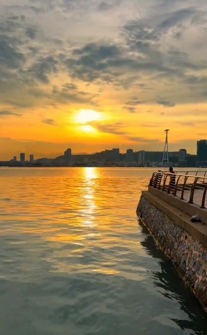 A scenic sunset over a body of water, with golden light reflecting on the waves, a city skyline in the distance, and a stone railing along a pier in the foreground.