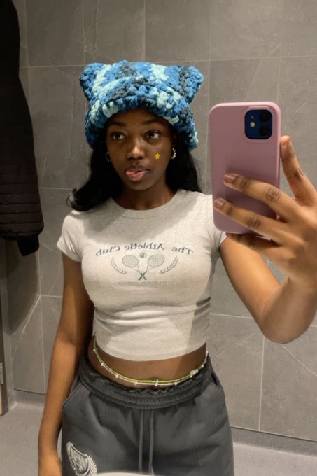 The image features a woman wearing a grey t-shirt with tennis rackets printed on it, holding up her phone to take a selfie. The background of the photo shows a bathroom setting with a mirror visible behind the woman. The caption at the bottom of the image reads "good morning live, love, laugh and mind your business stay hydrated, moisturized and keep playing magic fingers talk about $RIVER if you must and use Spenda to touch grass".