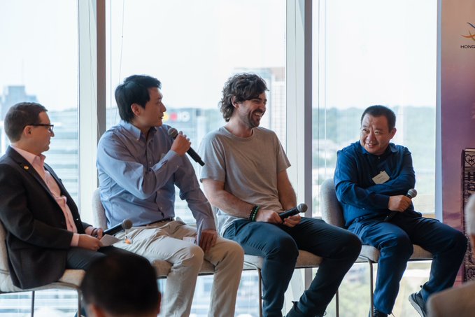 Four men sit on a panel discussion in a modern venue with large windows, two holding microphones, with Hong Kong signage partially visible in the upper right.