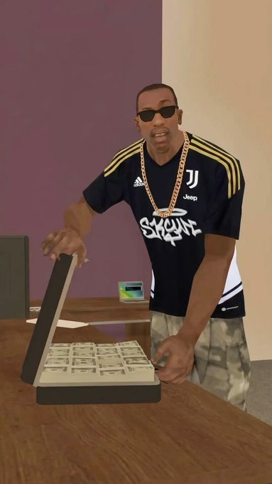 A man wearing sunglasses, gold chains, and a Juventus soccer jersey leans on a wooden desk next to an open briefcase filled with stacks of cash.