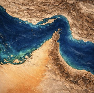 The image shows a painting depicting an underwater view of the Strait of Hormuz, with blue water surrounding the land mass. The painting features a river flowing through the area, surrounded by mountains and sand. In the background, there are reports about a new insurance service for shipping companies operating in the strait, which is aimed at providing coverage to ships during their passage through the strait of Hormuz.
