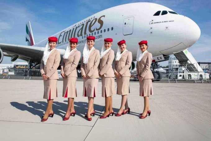 Six Emirates airline flight attendants in beige uniforms and red hats pose in front of an Airbus A380-800 aircraft on the tarmac.