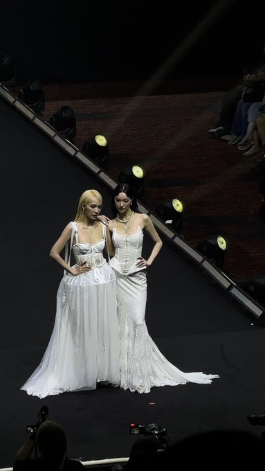 Two women in elaborate white gowns pose on a dimly lit stage with overhead spotlights.