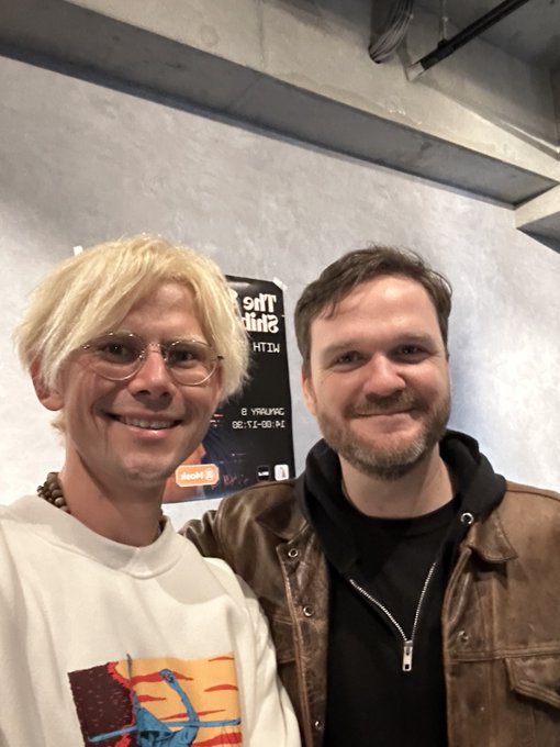 A selfie of two men smiling, one with blonde hair and glasses wearing a white shirt, the other with a beard wearing a brown jacket and black hoodie, with a poster behind them.