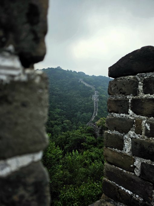 Atmospheric photo of the Great Wall of China viewed through a gap in ancient brick battlements, with misty forested mountains in the background.