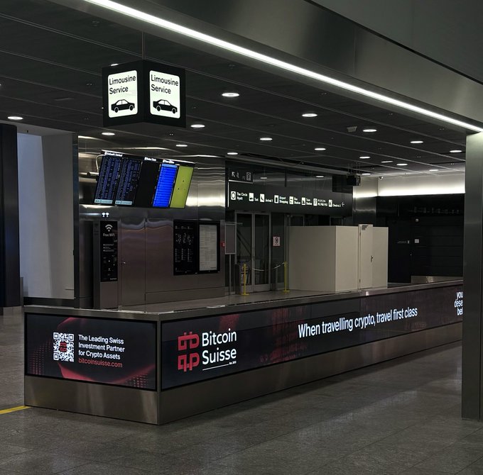 A "Bitcoin Suisse" branded kiosk advertising crypto services with the tagline "When travelling crypto, travel first class" and a "Limousine Service" sign above it, located in what appears to be an airport arrivals hall, directly supporting the post's observation about unexpectedly encountering the company upon landing in Zürich.