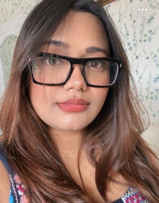 A close-up selfie of a young woman with long, wavy dark brown hair wearing black rectangular glasses and a floral patterned top, looking directly at the camera.