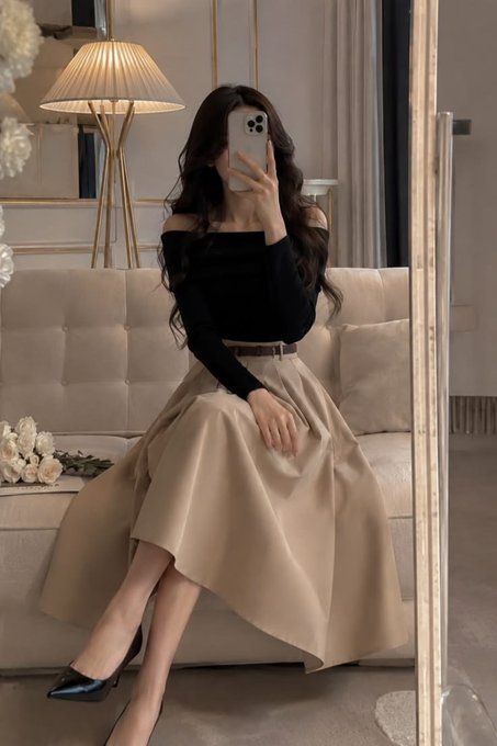 A woman taking a mirror selfie wearing a black off-shoulder top and a beige midi skirt with a brown belt, standing in a stylish room with a beige sofa.