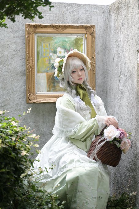 A young woman with long silver hair and a straw bonnet, dressed in a vintage-style light green and white dress, posing with a basket of roses in front of a gold-framed painting.