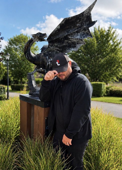 A man in a black hoodie and 'TDE' cap standing next to a dark dragon statue in an outdoor garden setting, holding his cap.