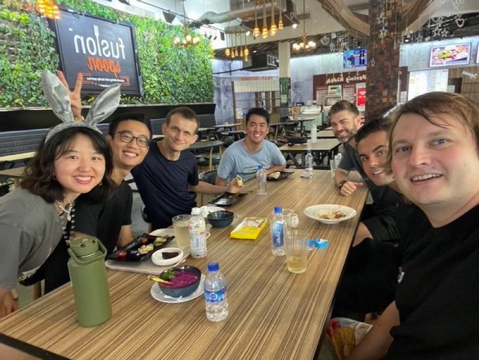 A group of eight colleagues gathers around a wooden table at a "nolefú noodle" restaurant, smiling for the camera with water bottles and dishes spread across the table, capturing a long-overdue team meal together.