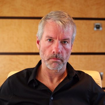 A middle-aged man with gray hair and a goatee, wearing a black button-down shirt, sits against a wooden paneled background with a serious expression. The image relates to the post as the man appears to be Michael Saylor, the prominent Bitcoin advocate known for his views on crypto protection and utility.