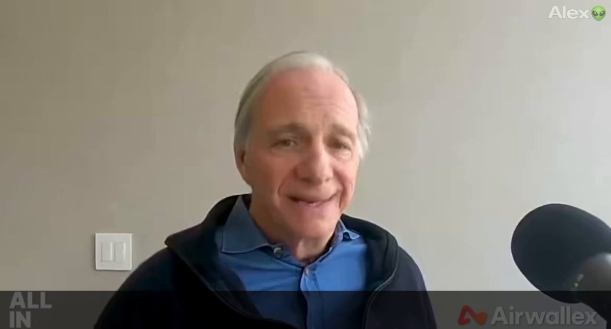 The image shows a man wearing a blue shirt, smiling at the camera while holding a microphone. The background of the image features a white wall with a clock hanging on it. There are also some text elements visible in the image that read "All In" and "Airwalx".