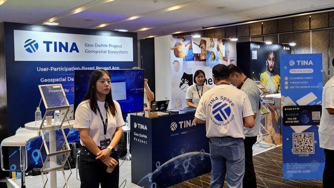 The image shows an event booth with a woman wearing a white shirt standing next to a man in jeans and another woman wearing a black t-shirt also standing near the booth, likely participating in or observing the event. The booth displays an advertisement for TINA (Tina's Atlas), and there are two laptops present on display as well. Additionally, people around the booth appear engaged with the content displayed at the event. A sign posted nearby indicates that the event has a reward system where individuals can earn points by participating or learning more about TINA's GeoDePIN distribution technology.