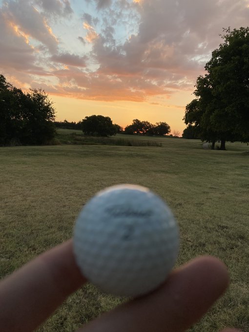 A hand holding a golf ball in the foreground with a scenic grassy field, trees, and a colorful sunset sky in the background.