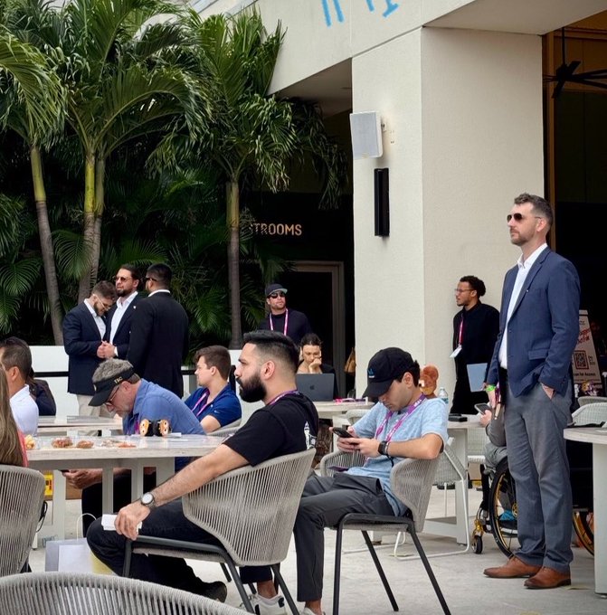 The image shows a group of people sitting at tables outside, possibly enjoying food or drinks. There are several chairs and dining tables scattered throughout the scene. A man wearing sunglasses stands near one of the tables, while another person can be seen using their cell phone. The backdrop features palm trees and a building with a sign that reads "Pull up for the Great Reset".