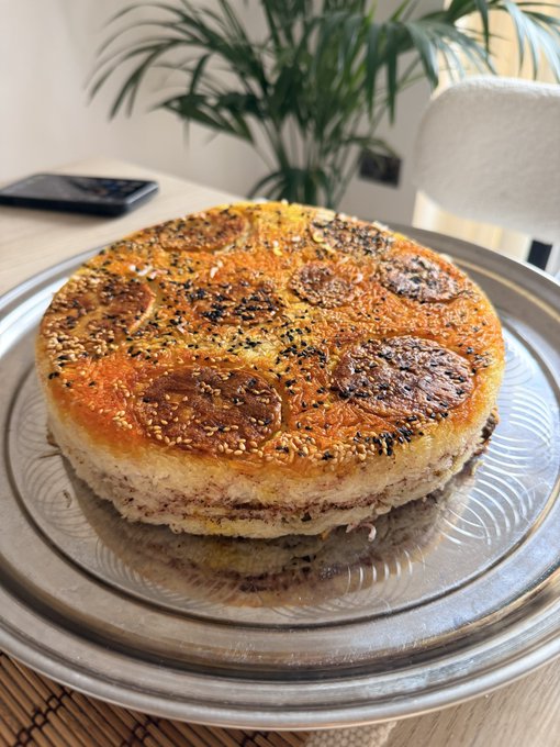 A round cake topped with sesame seeds and caramelized fruit slices sits on a silver tray, with a green potted plant in the background.
