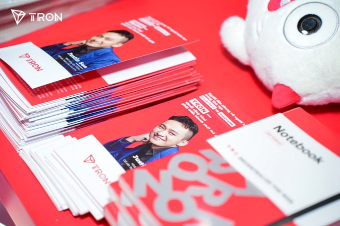 A stack of red TRON-branded promotional notebooks featuring a young man's photo, alongside a white plush toy and a 'NoteBook' card on a red surface.
