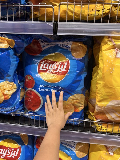 A child's hand reaches for a blue bag of Lay's Tomato Ketchup flavored potato chips on a store shelf, with yellow bags visible nearby.
