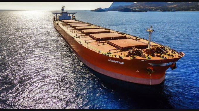An aerial view of a large red bulk carrier ship named 'GOODSHIP' sailing through bright blue ocean waters near mountainous coastlines.