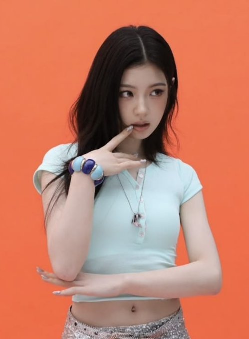 A young woman with long dark hair posing against an orange background, wearing a light blue cropped polo top and silver sequined skirt, with her hand raised near her chin.