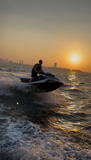 A jet ski rider carves through ocean waves at sunset, silhouetted against a glowing golden sky and a distant city skyline. The dramatic scene captures the essence of "GM" (Good Morning), greeting the new day with adventure on the open water.