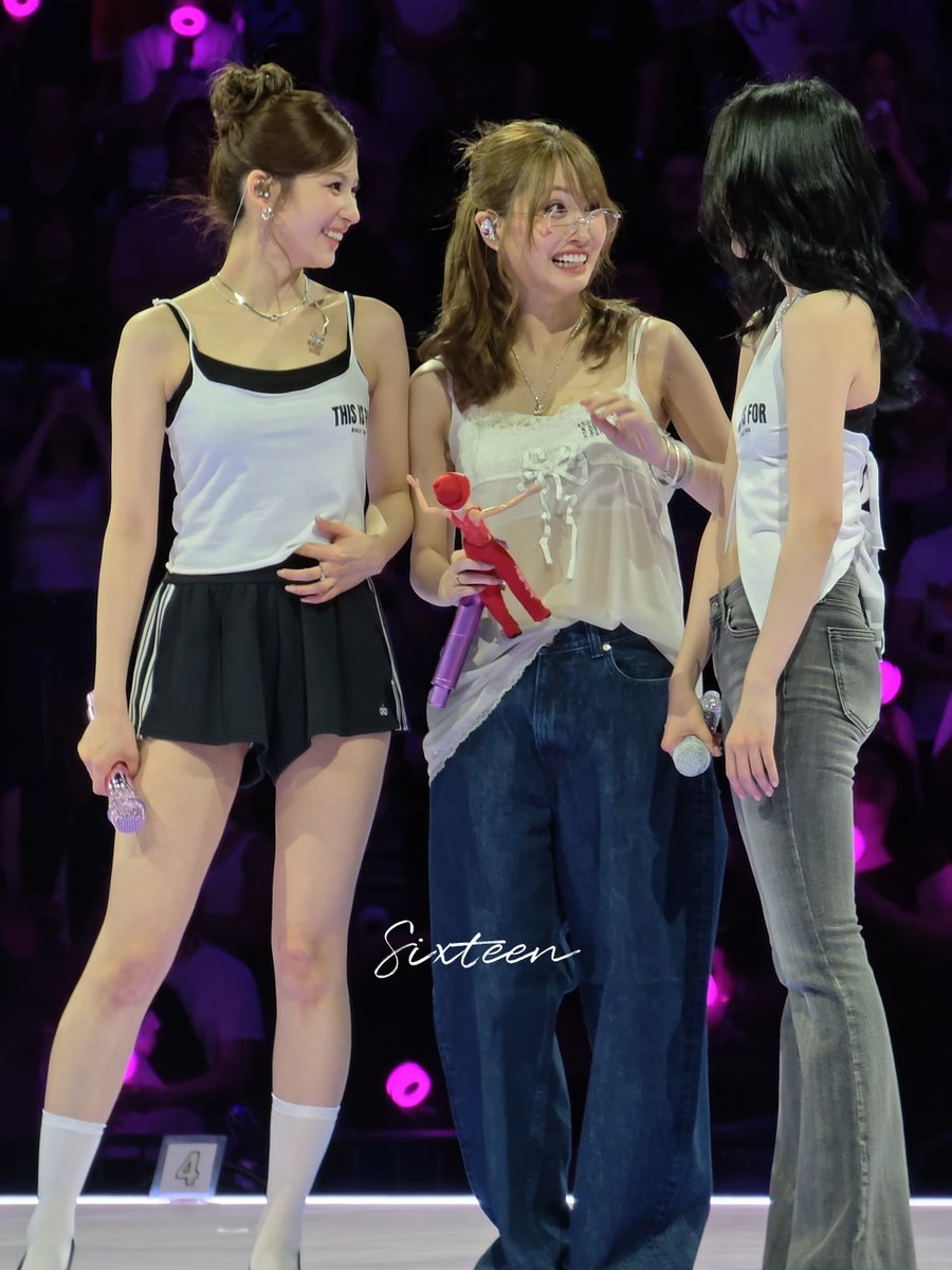 The image shows three women standing next to each other, possibly posing for a photo or participating in an event. One of them has her hair styled in pigtails, while another woman is wearing glasses. The third woman appears to be holding something in her hand. The background reveals that they are on stage with purple lights and a crowd of people watching the performance. A caption below the image reads: "What the fuck is wrong with mina".