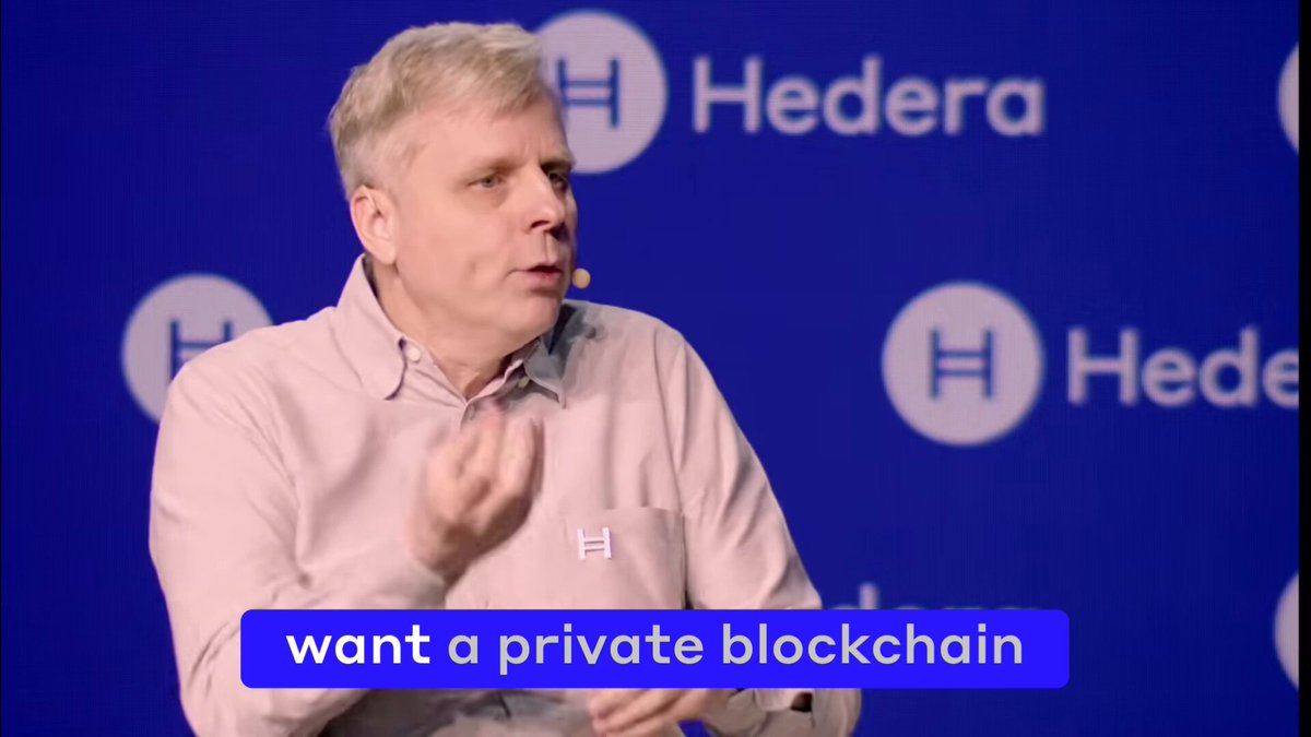 The image shows a man wearing a white shirt talking into a microphone, with "wants a private blockchain" written above him. The background features a blue wall with logos for "Hedera". This image may be used as part of an advertisement or promotional material related to blockchain technology and the concept of private blockchains.