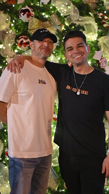 Two men pose smiling together in front of a decorated Christmas tree, one wearing a white 'adcc' t-shirt and cap, the other in a black 'DON'T FEED THE LOCALS' shirt.