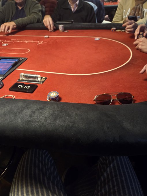 A first-person view from a poker table shows the edge of the red felt with chips and aviator sunglasses, while other players' hands and chips are visible across the table, with a sign reading 'TX-03'.