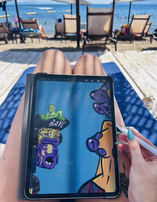 A first-person perspective of someone with turquoise nails drawing on a tablet with an Apple Pencil, viewing a cartoonish hip-hop style illustration with the word 'BAKU' visible; the scene is set at a beach with loungers and umbrellas in the background.
