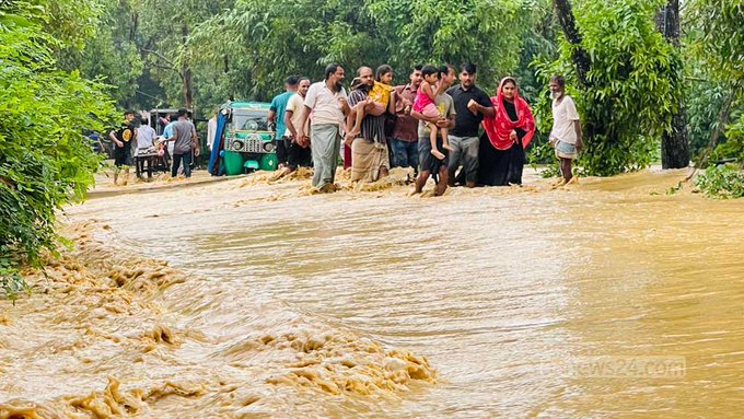 A group of people, including women in hijabs and children, walking through a flooded street with muddy brown water flowing across the road, with a green auto-rickshaw visible in the background.