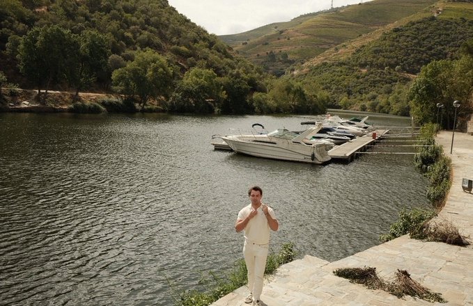 A man in a white outfit stands on stone steps by a lake, with boats docked nearby and forested hills in the background.