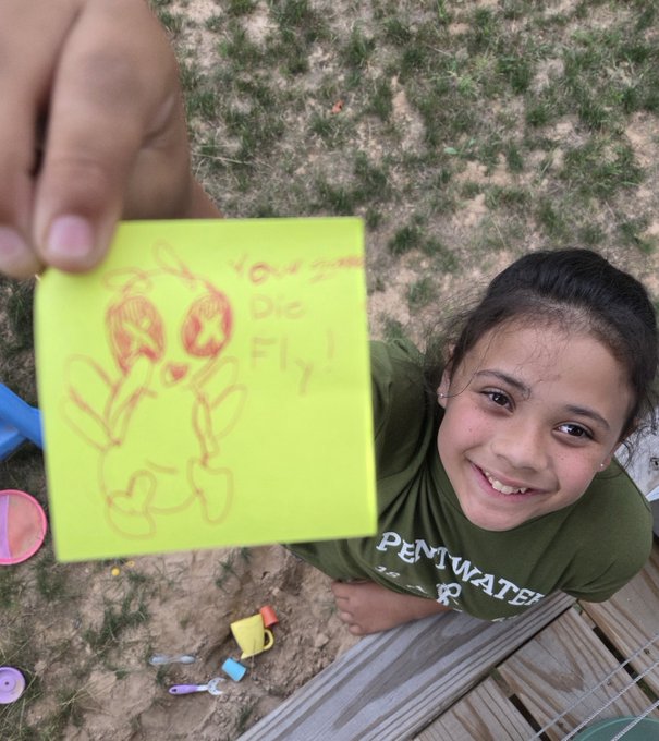 A smiling young girl wearing a green 'DEEPWATER' t-shirt holds up a yellow sticky note with a crude drawing and the handwritten text 'Never Die Fly'.