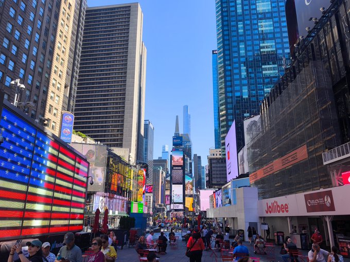 A bustling daytime scene of Times Square in New York City, featuring tall skyscrapers, bright digital billboards including an American flag display, and crowds of people, with a Jollibee storefront visible on the right.