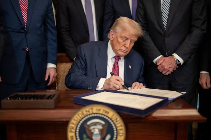 Donald Trump sits at a desk bearing the Presidential Seal, signing a document while wearing a dark suit and red tie, surrounded by several standing officials.