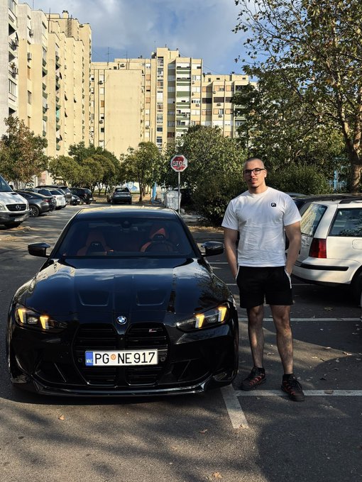 A man in a white t-shirt and black shorts stands next to a black BMW with a license plate reading 'PG NE 917' in a parking lot.