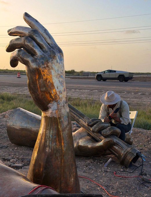 A scene showing large bronze-colored sculptures of hands and arms in a desert landscape, with a man in a cowboy hat working on one of the sculptures while a Tesla Cybertruck drives by in the background.
