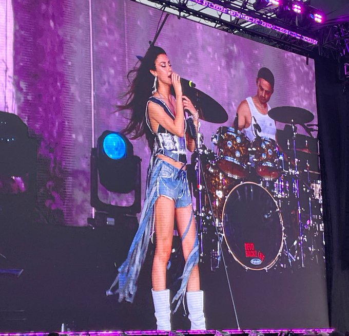 Female performer singing on stage in a silver and black outfit with denim shorts and flowing fabric, backed by a drummer on stage.