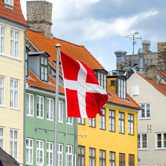The Danish flag waves prominently in front of colorful traditional townhouses, evoking Denmark's national identity. The image directly illustrates the post about Denmark's central bank concerns regarding foreign stablecoins' impact on the country's payments and monetary policy.