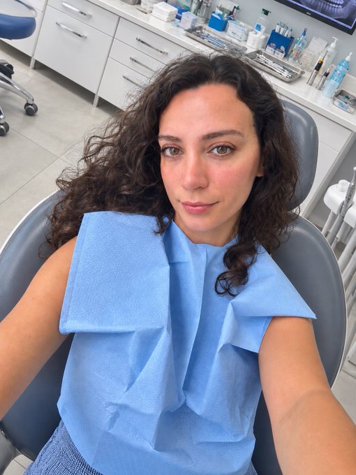 A selfie of a woman with curly dark hair sitting in a dental chair, wearing a blue protective bib in what appears to be a dentist's office.