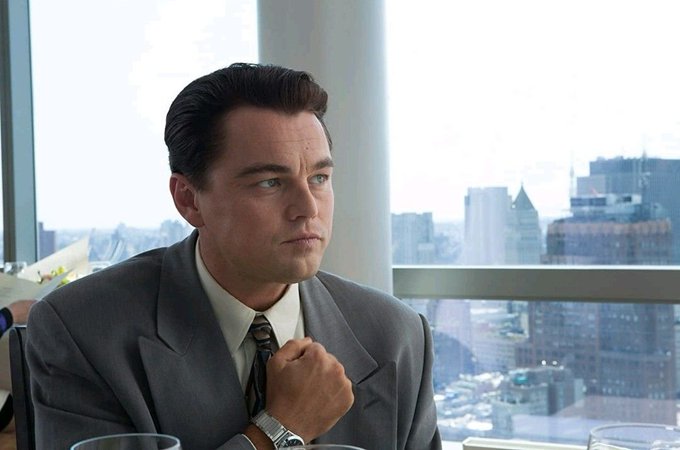 Leonardo DiCaprio in a gray suit, looking contemplative while adjusting his tie, with a city skyline visible in the background.