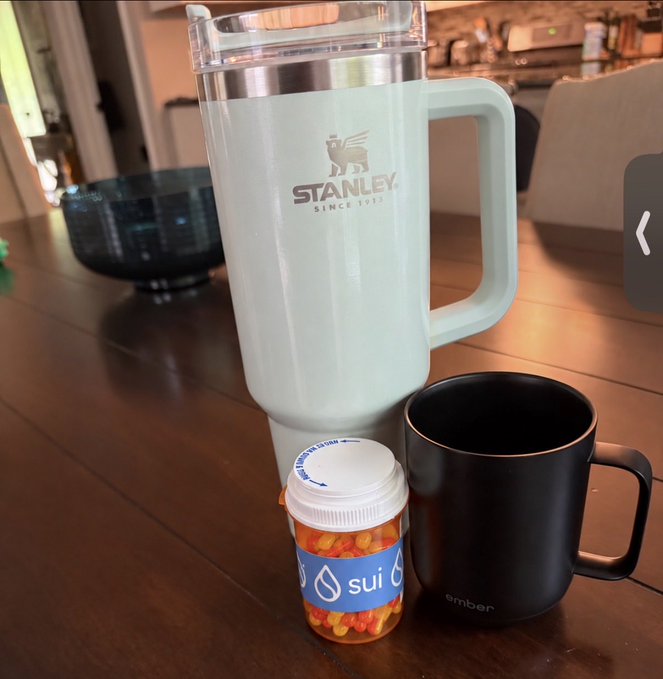 A mint green Stanley tumbler sits on a wooden table next to a small 'sui' branded pill bottle filled with orange pills and a black Ember mug.