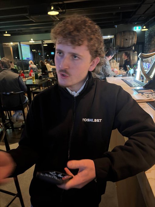 A young man wearing a black jacket with "TOSHI.BET" embroidered on it holds a phone in a bar setting with wooden barrels in the background, illustrating the caption's story about taking profits from a meme coin investment and turning them into a freewheeling lifestyle via crypto gambling.