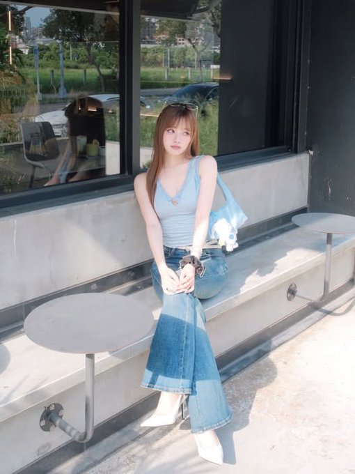 A young woman sits on a metal bench outside a building with dark windows, wearing a light blue ribbed tank top, jeans, and white pointed boots, holding a small bag and a plush toy.