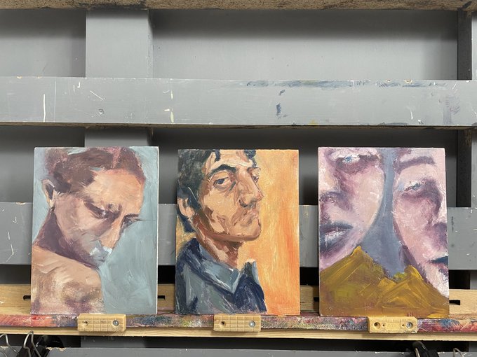 Three expressive oil painting portraits of men displayed side by side on a wooden easel or shelf against a grey background, each showcasing different color palettes and character studies.