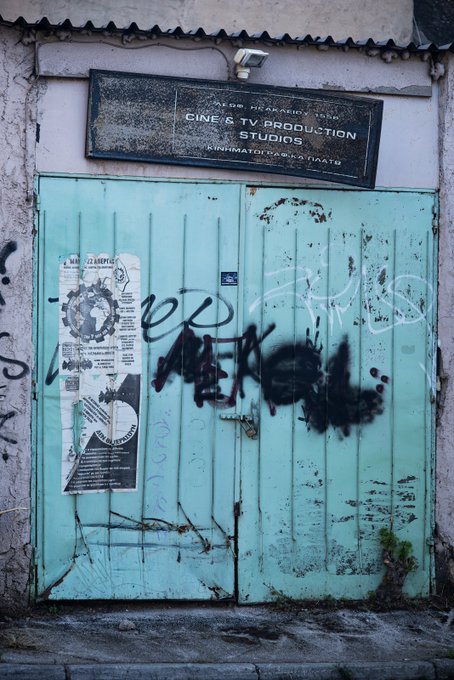 A photograph of a weathered turquoise metal door with graffiti and faded posters, located beneath a sign reading 'CINE & TV PRODUCTION STUDIOS' at 39 Melpomenis and Frynimatis streets.
