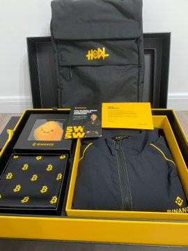 A Binance 9th anniversary gift box is displayed, featuring a "BN" branded backpack, folded Binance apparel with "BINANCE" and "B" logos, a yellow information card, and small packaged items. The unboxing reveals premium merchandise including clothing, a BTC-themed scarf, and a Binance AI pendant as mentioned in the post.